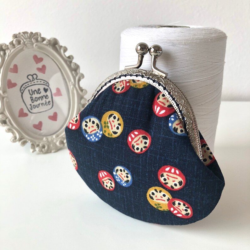 May include: A small, dark blue coin purse with a silver clasp. The purse is decorated with a repeating pattern of red, yellow, and blue Japanese Daruma dolls.