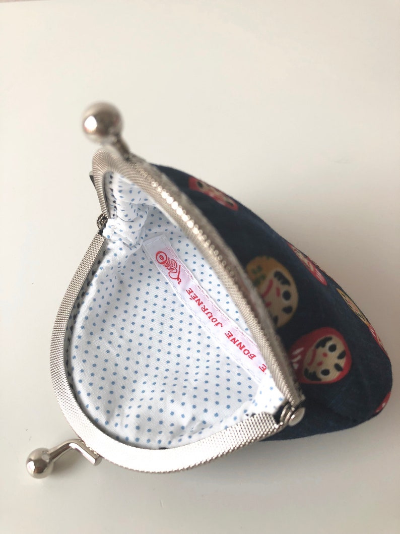 May include: A blue fabric coin purse with a silver clasp. The purse is decorated with a pattern of Japanese dolls. The inside of the purse is lined with white fabric with blue polka dots. The label inside the purse reads "Bonne Journee".