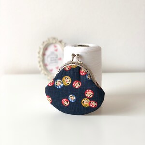 May include: A small, blue coin purse with a silver clasp. The purse is decorated with a repeating pattern of red and yellow Japanese daruma dolls.