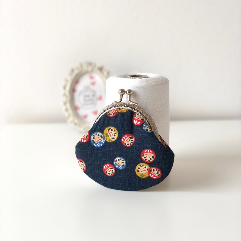 May include: A small, blue coin purse with a silver clasp. The purse is decorated with a repeating pattern of red and yellow Japanese daruma dolls.