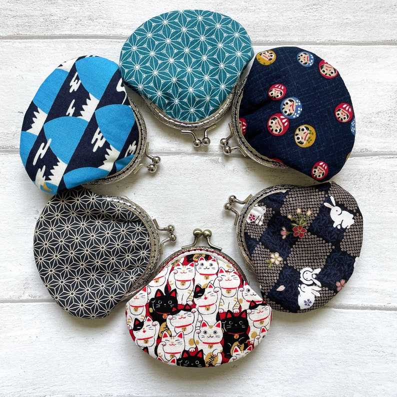 May include: Six different coin purses with various Japanese-inspired patterns. The purses are made of fabric and have metal frames. The patterns include a blue and white geometric design, a green and white geometric design, a black and white geometric design, a black and white floral design, a black and white cat design, and a blue and white design with red and gold accents.