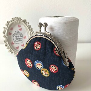 May include: A small, dark blue coin purse with a silver clasp. The purse is decorated with a repeating pattern of colorful Japanese Daruma dolls.