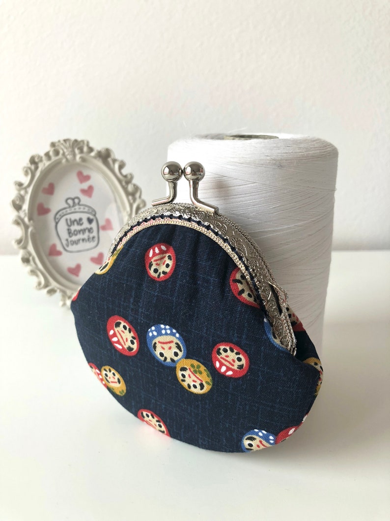 May include: A small, dark blue coin purse with a silver clasp. The purse is decorated with a repeating pattern of colorful Japanese Daruma dolls.