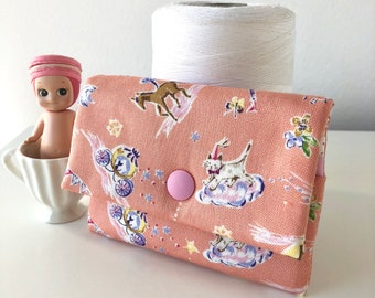 Puede incluir: Una pequeña cartera de monedas de tela rosa con un diseño caprichoso que presenta caballos, gatos y estrellas. La cartera tiene un cierre de botón y está hecha de una tela suave de algodón.