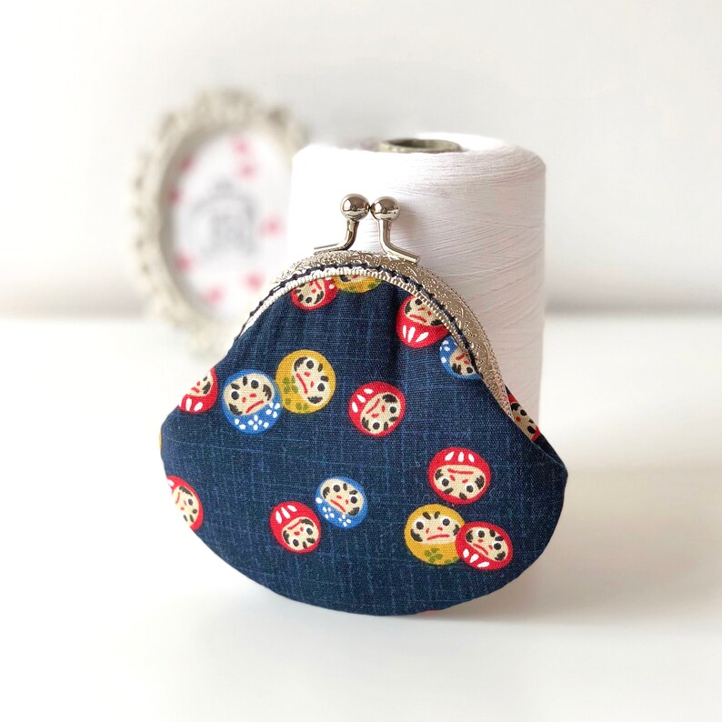 May include: A small, blue fabric coin purse with a silver clasp. The purse is decorated with a repeating pattern of red, yellow, and blue Japanese daruma dolls.