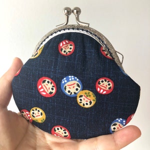 May include: A small, dark blue coin purse with a silver clasp and a white trim. The purse is decorated with a repeating pattern of colorful Japanese daruma dolls.