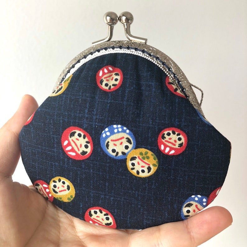 May include: A small, dark blue coin purse with a silver clasp and a white trim. The purse is decorated with a repeating pattern of colorful Japanese daruma dolls.
