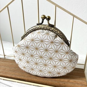 Peut inclure: Un porte-monnaie blanc avec un motif géométrique doré. Le sac à main a un fermoir de style vintage avec une finition bronze. Le sac à main est posé sur une étagère en bois.