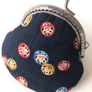 May include: A small, dark blue coin purse with a silver clasp and a silver handle. The purse is decorated with a repeating pattern of colorful Japanese daruma dolls.