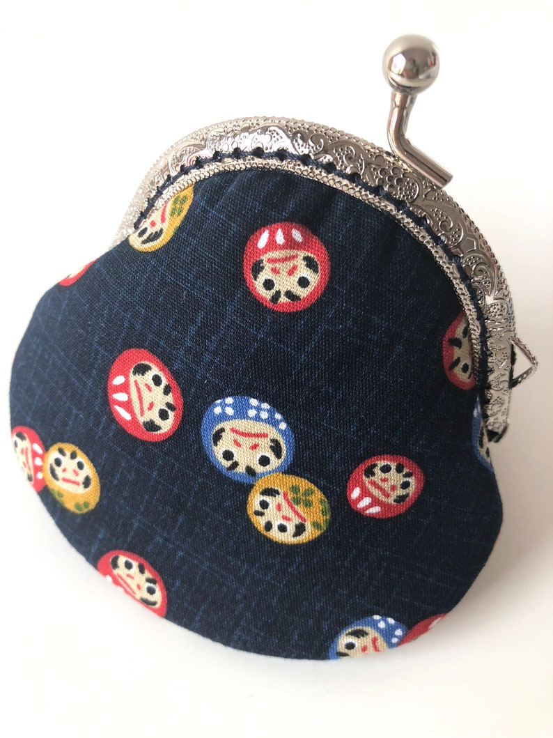 May include: A small, dark blue coin purse with a silver clasp and a silver handle. The purse is decorated with a repeating pattern of colorful Japanese daruma dolls.