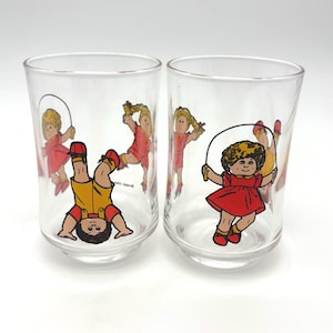 May include: Two clear glass tumblers with cartoon illustrations of children playing. One tumbler features a child in a yellow outfit doing a handstand. The other tumbler has a child in a red dress jumping rope. The glasses are on a white background.
