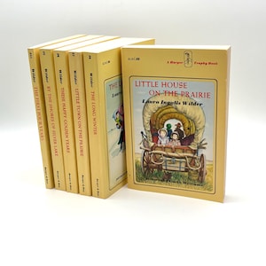 May include: A collection of vintage children's books, featuring titles like "Little House on the Prairie" and "The Long Winter." The books have a cream-colored cover with illustrations and text. The spines display the author's name and book titles.