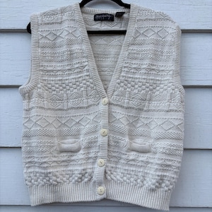 May include: Cream-colored knit vest with a V-neck, two front pockets, and button closure. The vest features a textured pattern throughout and is hanging on a black hanger against a light blue background.