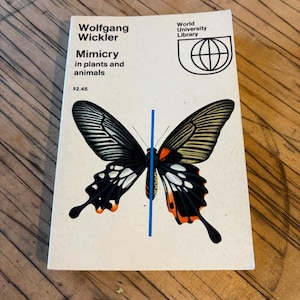 May include: A vintage book titled "Mimicry in plants and animals" by Wolfgang Wickler. The cover features a detailed illustration of a butterfly with black, white, orange, and yellow wings. The book has a white cover with a blue vertical line.
