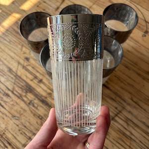 Puede incluir: Un juego de seis vasos altos vintage. El vidrio transparente presenta líneas verticales grabadas, y el borde superior es plateado con un diseño floral ornamentado. Los vasos están dispuestos sobre una superficie de madera.