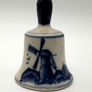 May include: A ceramic bell with a blue and white windmill design. The bell features a dark blue handle and a detailed illustration of a windmill, clouds, and birds. The bell is a decorative item.