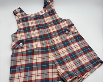 May include: A sleeveless romper featuring a plaid pattern in red, white, blue, and yellow. The garment has two shoulder straps with white buttons, two side pockets with buttoned flaps, and a snap closure at the bottom hem for easy access.