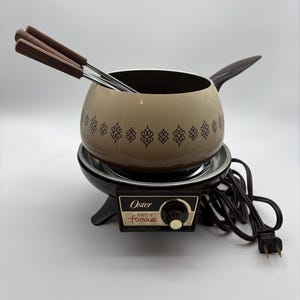 May include: An Oster electric fondue pot with a beige bowl featuring a brown geometric pattern, a dark brown handle, and a black base with a temperature control dial. Three metal fondue forks with brown wooden handles are placed inside the pot.