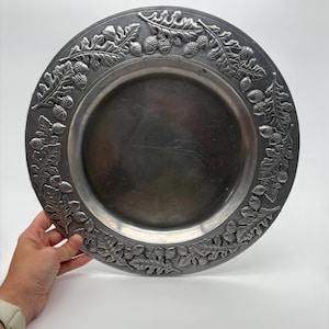 May include: A round, silver-toned metal plate with a decorative border of oak leaves and acorns. The plate has a slightly concave center and a wide, detailed rim. The plate is held by a hand with a white watch.