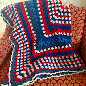 May include: A handmade crochet blanket in a granny square pattern. The blanket features a color scheme of red, white, and blue squares, with a white scalloped border. The blanket is draped over a red floral patterned chair.