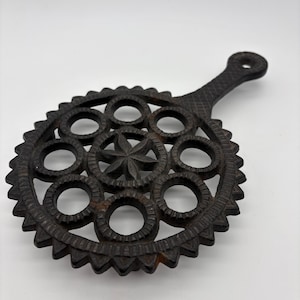 May include: A dark brown cast iron trivet with a handle. The trivet has a circular design with a star in the center and several circular openings. The outer edge has a decorative, pointed pattern. This kitchenware item is designed to protect surfaces from hot cookware.