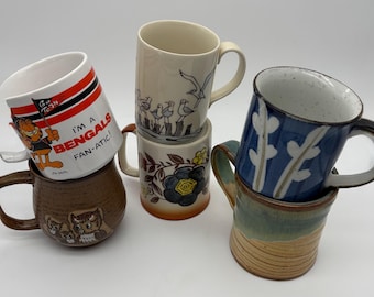 May include: A collection of six ceramic mugs stacked on top of each other. The mugs feature various designs, including cartoon characters, floral patterns, and bird illustrations. One mug has the text "I'm a Bengals Fan-atic!"