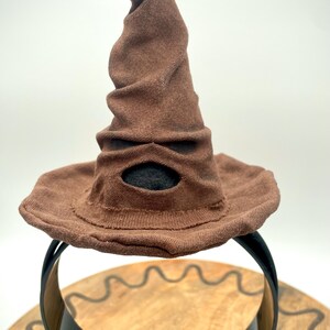 May include: A brown, wizard-style hat with a wide brim and a pointed, twisted top. The hat has a dark oval opening and is displayed on a black stand. The hat is made of a soft, textured material.