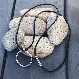 May include: A black leather cord necklace with a silver circle pendant. The necklace is lying on a pile of gray rocks.
