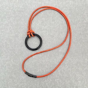 May include: A necklace featuring a thin, orange cord that loops through a smooth, black circular pendant. The cord is secured with a black metal clasp at the bottom, creating a continuous, adjustable loop design for this piece of jewelry.