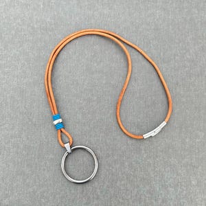 May include: A tan leather lanyard with a silver metal ring and clasp. The lanyard has a blue and silver accent near the ring. The background is a solid gray color.