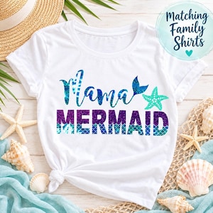 May include: A white short-sleeve t-shirt featuring the text Mama MERMAID in blue and purple glittery script and block lettering. The design includes a blue mermaid tail and a teal starfish. The text Matching Family Shirts is visible.