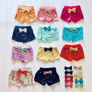 May include: Ten pairs of sequined shorts for girls, each with a different color and bow. The colors include pink, red, blue, green, gold, silver, and black.