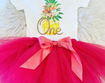 May include: A white onesie with a pineapple design and the word "One" in gold glitter. The onesie is paired with a pink tulle tutu and a gold glitter bow.