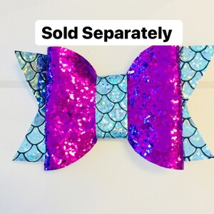 May include: A purple and blue glitter bow with a light blue mermaid scale pattern. The bow is tied in a knot and has a white background. "Sold Separately" is written in black text at the top of the image.