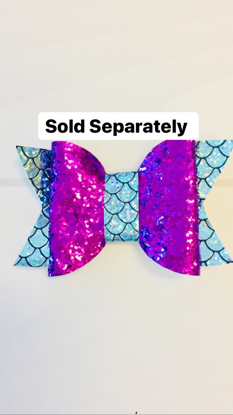 May include: A purple and blue glitter bow with a light blue mermaid scale pattern. The bow is tied in a knot and has a white background. "Sold Separately" is written in black text at the top of the image.