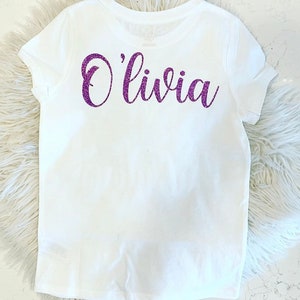 K&ouml;nnte beinhalten: Wei&szlig;es T-Shirt mit dem Namen "Olivia" in lila Glitzer auf der R&uuml;ckseite.