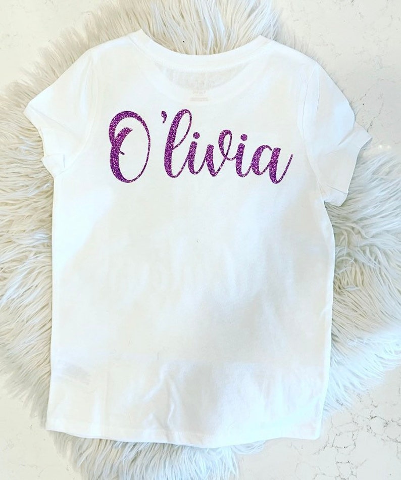 K&ouml;nnte beinhalten: Wei&szlig;es T-Shirt mit dem Namen "Olivia" in lila Glitzer auf der R&uuml;ckseite.