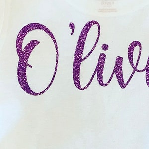 K&ouml;nnte beinhalten: Ein wei&szlig;es T-Shirt mit dem Wort "Olivia" in lila Glitzer.