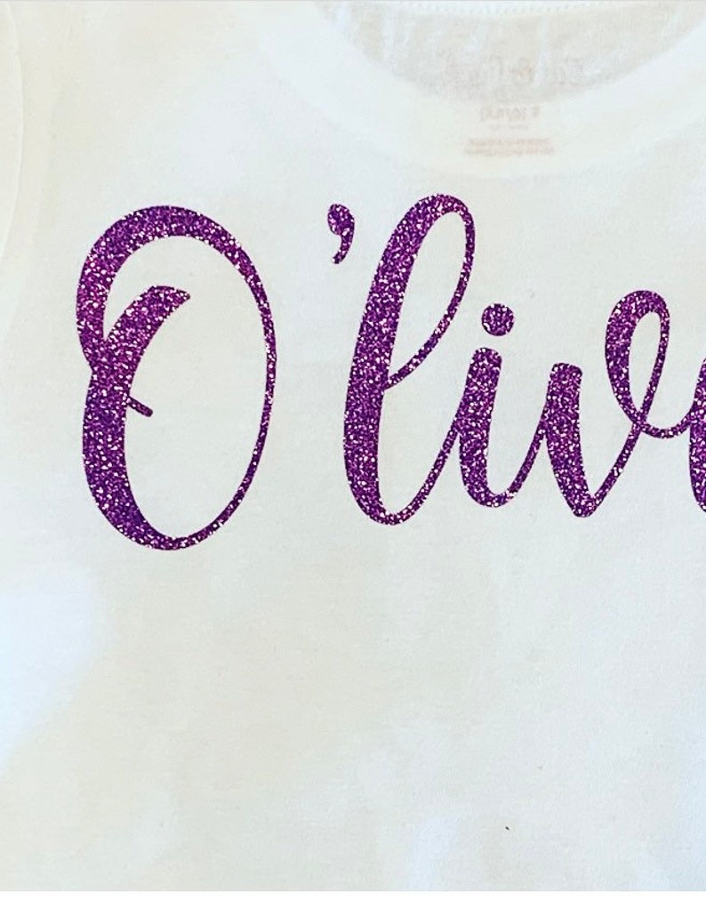 K&ouml;nnte beinhalten: Ein wei&szlig;es T-Shirt mit dem Wort "Olivia" in lila Glitzer.