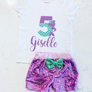 May include: A white t-shirt with a purple number 5 decorated with green glitter scales and a mermaid. The shirt also has the name "Giselle" written in purple glitter. The shirt is paired with purple sequin shorts with a green bow.