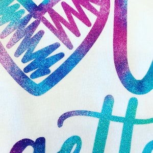K&ouml;nnte beinhalten: Ein wei&szlig;es T-Shirt mit einem glitzernden blau-violetten Herzen und dem Wort "gotta" in glitzerndem Blau und Lila.