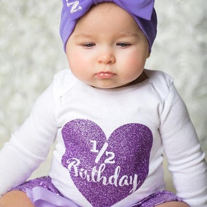May include: A baby girl wearing a white onesie with a purple glitter heart that says "1/2 Birthday". She is also wearing a purple headband with a bow and the word "Queen" on it. She is wearing a purple sequined skirt with a matching purple bow.