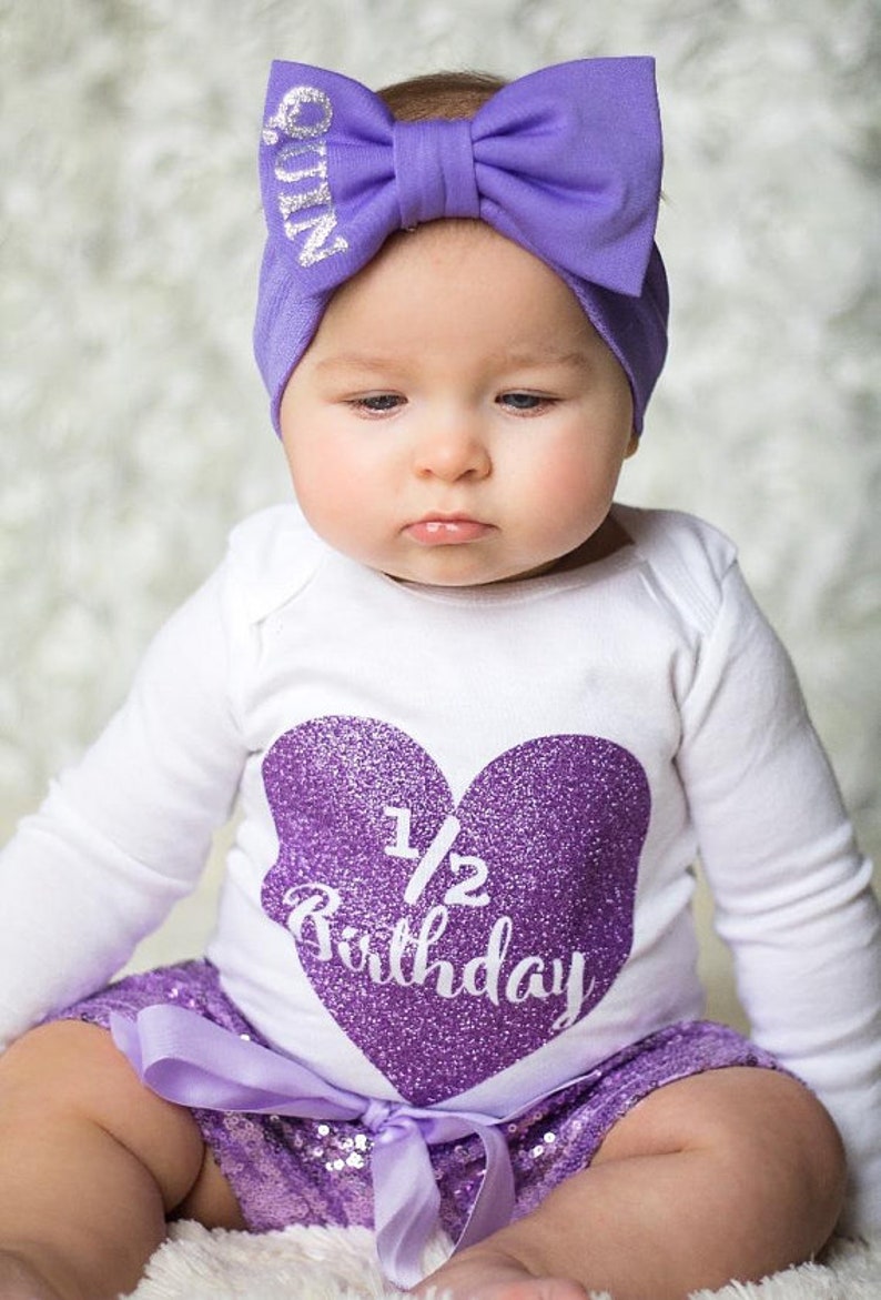 May include: A baby girl wearing a white onesie with a purple glitter heart that says "1/2 Birthday". She is also wearing a purple headband with a bow and the word "Queen" on it. She is wearing a purple sequined skirt with a matching purple bow.