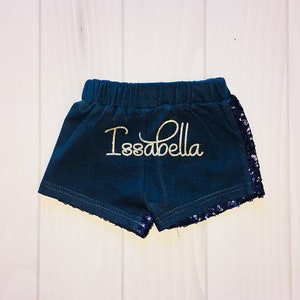 May include: Dark blue shorts with a gold glitter "Isabella" name on the front. The shorts have a black sequin trim around the bottom.