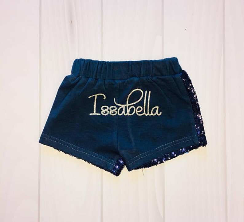 May include: Dark blue shorts with a gold glitter "Isabella" name on the front. The shorts have a black sequin trim around the bottom.