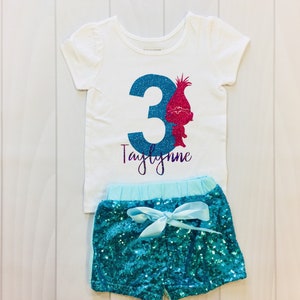 May include: A white t-shirt with a blue number 3 and a pink troll character. The shirt says "Taylynne" in purple glitter. The shirt is paired with turquoise sequin shorts with a light blue waistband and a light blue bow.