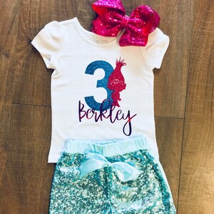 May include: A white t-shirt with a blue number 3 and a pink glitter troll character. The shirt says "Berkeley" in purple glitter. The shirt is paired with turquoise sequin shorts and a pink bow.