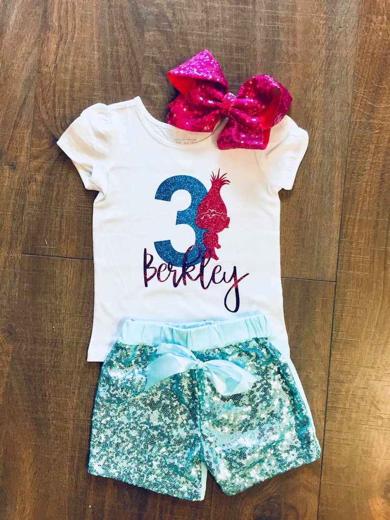 May include: A white t-shirt with a blue number 3 and a pink glitter troll character. The shirt says "Berkeley" in purple glitter. The shirt is paired with turquoise sequin shorts and a pink bow.