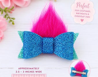 May include: A blue glitter hair bow featuring a vibrant hot pink faux fur tuft on top. The bow measures approximately 2.5 to 3 inches wide as shown by a ruler. The back includes a metal alligator clip for attachment to hair or accessories.