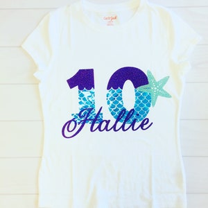May include: White t-shirt with a purple and blue glitter number 10 with mermaid scales and a starfish. The name "Hallie" is written in purple glitter below the number.